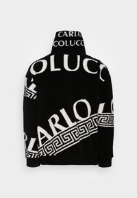 Black fleece jacket with large white lettering and geometric patterns. High collar, relaxed fit. Textured fabric, visible seams, no fasteners.