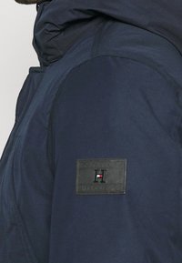 Marinblå jacka med en texturerad yta, prydd med en svart läderpatch med logotypen 'TOMMY HILFIGER'. Designen inkluderar en huva och en strömlinjeformad silhuett.