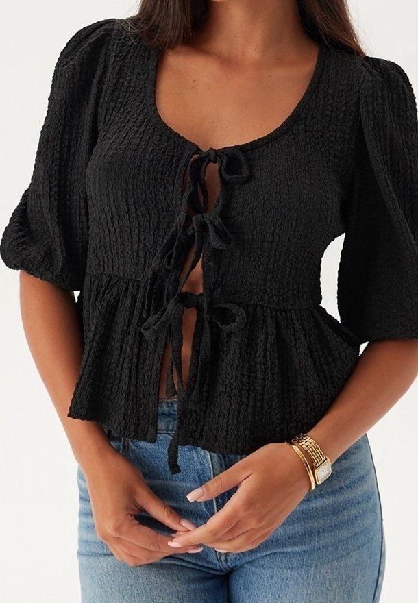 Femme portant un chemisier noir texturé avec des manches bouffantes et trois liens de fermeture à l'avant, associé à un jean bleu et un bracelet de montre en or.