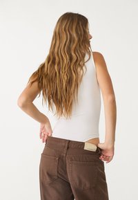 Witte mouwloze bodysuit gecombineerd met hooggetailleerde bruine jeans. De outfit heeft een gestikte label aan de tailleband en textiel met textuur.