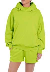Felpa verde brillante con cappuccio e pantaloni corti abbinati realizzati in tessuto testurizzato. La felpa presenta una tasca a marsupio e un dettaglio del logo sul petto.