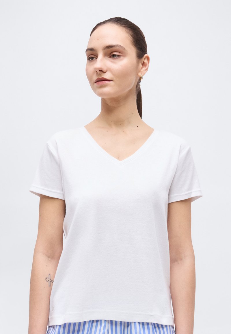 Γυναίκα με τα μαλλιά πιασμένα πίσω, φορώντας λευκό μπλουζάκι με V λαιμόκοψη και κοντά μανίκια και μπλε παντελόνι με ρίγες, γυρισμένη ελαφρώς προς τα αριστερά σε απλό φόντο.