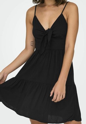 Femme portant une robe noire sans manches avec de fines bretelles, un nœud à l'avant et une jupe froncée, tenant l'ourlet d'une main.