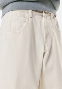 Beige denimbukser med lige snit. Har to forlommer og synlige syninger. Stoffet har en glat tekstur.