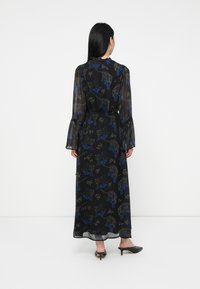 Kvinna med långt svart hår iförd en svart maxiklänning med blåa och grå blommönster och genomskinliga långa ärmar, står med ryggen mot kameran.