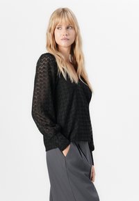 Kvinna med långt blont hår som bär en svart texturerad blus med långa ärmar och grå veckade byxor, ser åt vänster med en hand i fickan.