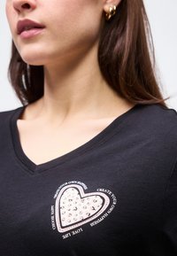 Γυναίκα που φοράει μαύρο μπλουζάκι με λαιμόκοψη V, διακοσμημένο με καρδιά από τρουκς και κείμενο "Δημιούργησε τη δική σου ευτυχία, αγάπα τη ζωή, επίλεξε να είσαι χαρούμενος" στο στήθος.