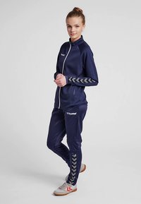 Granatowy dres wykonany z gładkiej tkaniny, z białym zamkiem błyskawicznym, logo oraz szarymi wzorami w kształcie łyżew na rękawach i spodniach. Jasnobrązowe trampki.