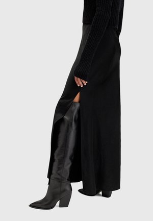 Femme portant une longue jupe noire fendue sur le côté et des bottes en cuir noires à talons montant jusqu'aux genoux, debout contre un fond uni.