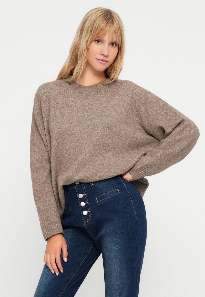 Jasnobrązowy sweter dziany z okrągłym dekoltem i opadającymi ramionami, w zestawieniu z ciemnoniebieskimi spodniami z wysokim stanem z metalowymi guzikami.