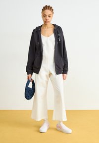 Sudadera con cremallera de color gris oscuro con bolsillos, mono sin mangas blanco, bolso tejido de color azul marino y zapatos de slip-on rosa con suelas blancas. Fondo brillante.
