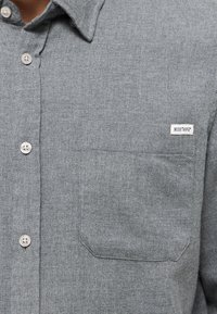 Graues Hemd mit Knopfleiste aus strukturiertem Stoff, ausgestattet mit einer Brusttasche und einem kleinen Markenlabel an der Seite. Die Knöpfe sind hell gefärbt.