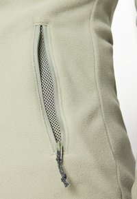 Vetoketjullinen fleece-takki vaaleanvihreänä, jossa on verkkosisältöinen tasku ilmanvaihtoa varten ja kiristysnauha säädettävää sulkemista varten.