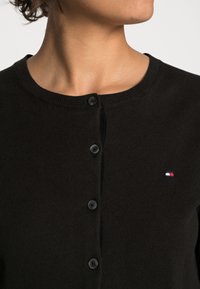Cardigan noir en tissu tricoté doux, avec un col rond, cinq boutons noirs et un petit logo en rouge, blanc et bleu.