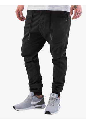 Mann trägt schwarze Jogginghose und graue Nike-Sneaker mit weißem Swoosh-Logo, steht vor einem schlichten weißen Hintergrund.