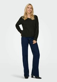 Maglione nero lavorato a maglia abbinato a jeans a zampa d'elefante blu scuro e scarpe col tacco nere. La modella sta in piedi con le mani sui fianchi, su uno sfondo neutro.