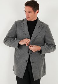 Sivi plašč iz volnene mešanice z ovratnikom z režo, spredaj zapenjan na gumbe, z dvema bočnima žepoma in krojenim pasom. Tkanina z izrazito strukturo.