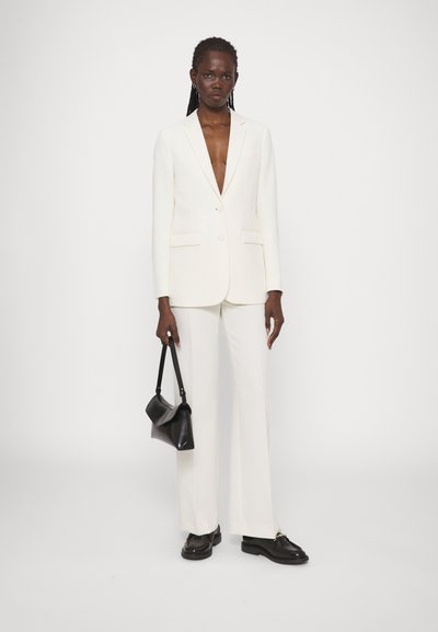Blazer bianco su misura con scollo a V profondo, pantaloni bianchi abbinati e una borsa nera strutturata. Tessuto elegante e silhouette moderna.