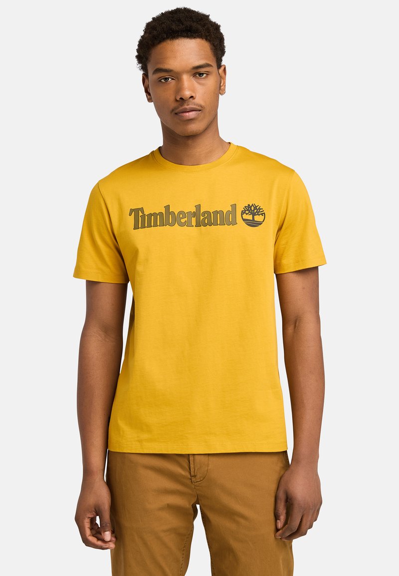 Keltainen puuvillapaita, jossa on tummahkon harmaa "Timberland" -logo ja puugrafiikka. Lyhyet hihat ja pyöreä pääntie.