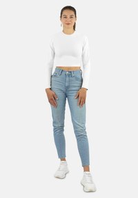 Weißes, langärmeliges, kurzes Oberteil, kombiniert mit hochtaillierten, hellblauen Jeans und weißen Sneakers, vor einem schlichten Hintergrund.