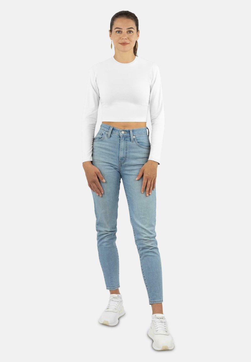 Weißes, langärmeliges, kurzes Oberteil, kombiniert mit hochtaillierten, hellblauen Jeans und weißen Sneakers, vor einem schlichten Hintergrund.