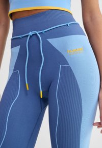 Blå idrottsleggings med olika nyanser av blått och ribbad textur, med dragsko och gula accenter nära midjan.