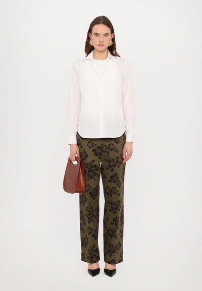 Chemise blanche à manches longues à boutons, pantalon imprimé floral vert olive, chaussures noires à bout pointu, tenant un petit sac à main marron.