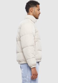 Jachetă puf light beige cu guler înalt, model matlasat, manșete elastice și buzunare laterale, purtată peste pantaloni din denim deschis.