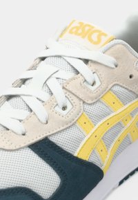 Asics sneakers med grå mesh-overdel, suède detaljer i beige og marineblå, samt gule sidestriper. Har hvite lisser og en polstret tunge.