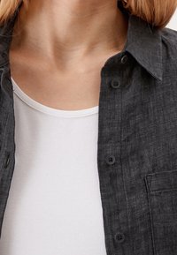 Chemise boutonnée gris foncé en tissu texturé, dotée d'un col et d'une poche poitrine, portée sur un t-shirt blanc à col rond.