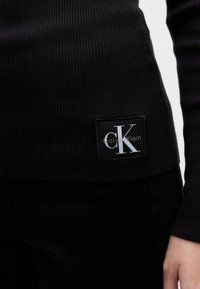 Fekete bordázott kötött hosszú ujjú felső, amelynek az alján egy jól látható Calvin Klein logó folt található. Texturált anyag, karcsú szabással.