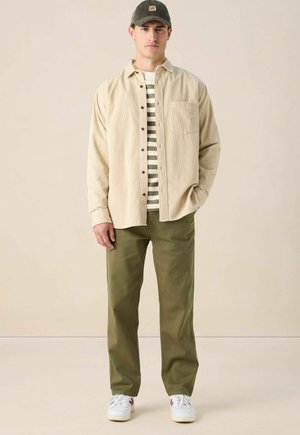 Beige ribfluwelen overhemd met knopen over een gestreept groen en wit T-shirt, gedragen met groene broek en witte sneakers met bordeauxrode accenten.