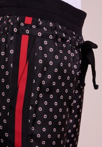 Svarta byxor med vita och röda blommönster, röda och svarta sidränder, elastisk midja och ett svart snöre knutet som en båge.