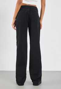 Femme portant un jean noir taille haute à jambes larges et un top blanc court, debout dos à la caméra sur un fond uni.