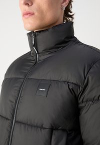 Svart puffjakke med høy krage, frontglidelås, kviltet tekstur, og Calvin Klein logo-patch på brystet.