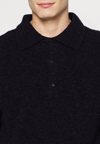 Dunkelblaues Woll-Polohemd mit einem breiten Kragen, vier Knöpfen und weicher Textur. Der Stoff scheint leicht strukturiert und warm zu sein.