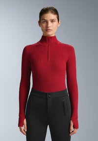Röd ribbad långärmad topp med hög krage och kvartszip. Tyget är texturerat med vertikala mönster, kombinerat med svarta figurnära byxor.