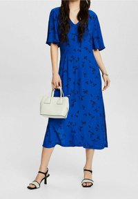 Robe midi bleue à imprimé floral avec des manches courtes évasées ; associée à un sac à main blanc et des sandales à talons noirs, mettant en avant un style décontracté.