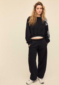 Sort cropped sweatshirt med tekstureret stof og hvid tekst på venstre ærme, kombineret med sorte sweatpants og hvide sneakers.