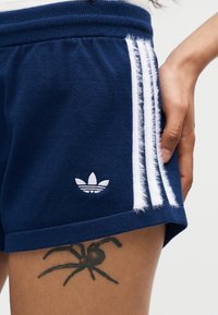 Κοντινό πλάνο σκούρων μπλε σόρτς Adidas με λευκές ρίγες, λογότυπο Adidas, τατουάζ αράχνης στο μηρό και ένα χέρι ακουμπισμένο στη μέση.