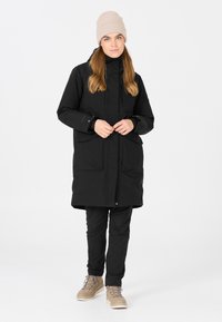 Svart vattentät parkas med framfickor, i kombination med svarta byxor och bruna stövlar. Modellen bär en ljusbeige stickad mössa.
