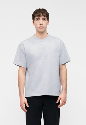 Jeune homme aux cheveux bruns bouclés portant un t-shirt ample à manches courtes gris clair et un pantalon noir sur un fond blanc uni.