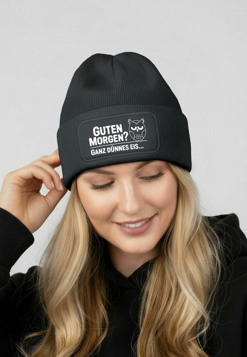 Black knit beanie featuring a rectangular patch with white text and a cartoon owl design. The fabric has a ribbed texture.