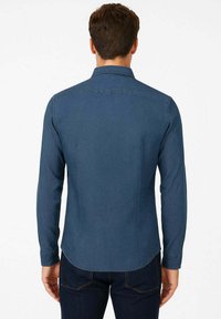 Camicia blu scuro a maniche lunghe con collo a punta, orlo posteriore e dettagli delle cuciture sul retro. Realizzata in un tessuto liscio simile al denim.