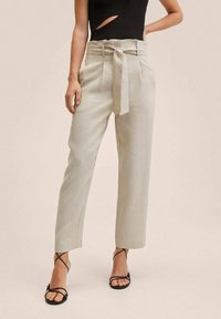 Jasno beżowe spodnie z wysokim stanem, z paskiem wiązanym, w strukturalnym materiale, o prostych nogawkach i bocznych rozcięciach, zestawione z czarnymi sandałami na paski.