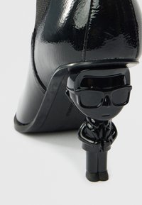 Μαύρο παπούτσι με ψηλό τακούνι από δερμάτινο δερμάτινο βινύλιο, με διακοσμητική μαύρη φιγούρα ανθρώπου με γυαλιά ως τακούνι. Λεία υφή, κομψός σχεδιασμός.