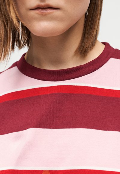 Zbliżenie osoby noszącej sweter w paski z poziomymi pasami w kolorach bordowym, czerwonym, różowym i białym, pokazujące tylko usta i szyję.