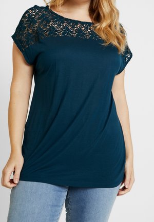 Vrouw die een donker petrolkleurig T-shirt met korte mouwen en bloemige kanten details bij de halslijn draagt, en lichtblauwe spijkerbroek, tegen een effen achtergrond.
