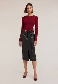 Femme portant un pull bordeaux ajusté avec des manches découpées, une jupe midi en cuir noir avec fente avant, une ceinture assortie et des talons hauts bordeaux.