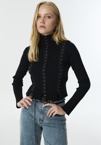 Haut noir à col roulé côtelé avec un design ajusté, boutons sur les côtés et clous argentés à l'avant. Associé à un Jean taille haute bleu clair.